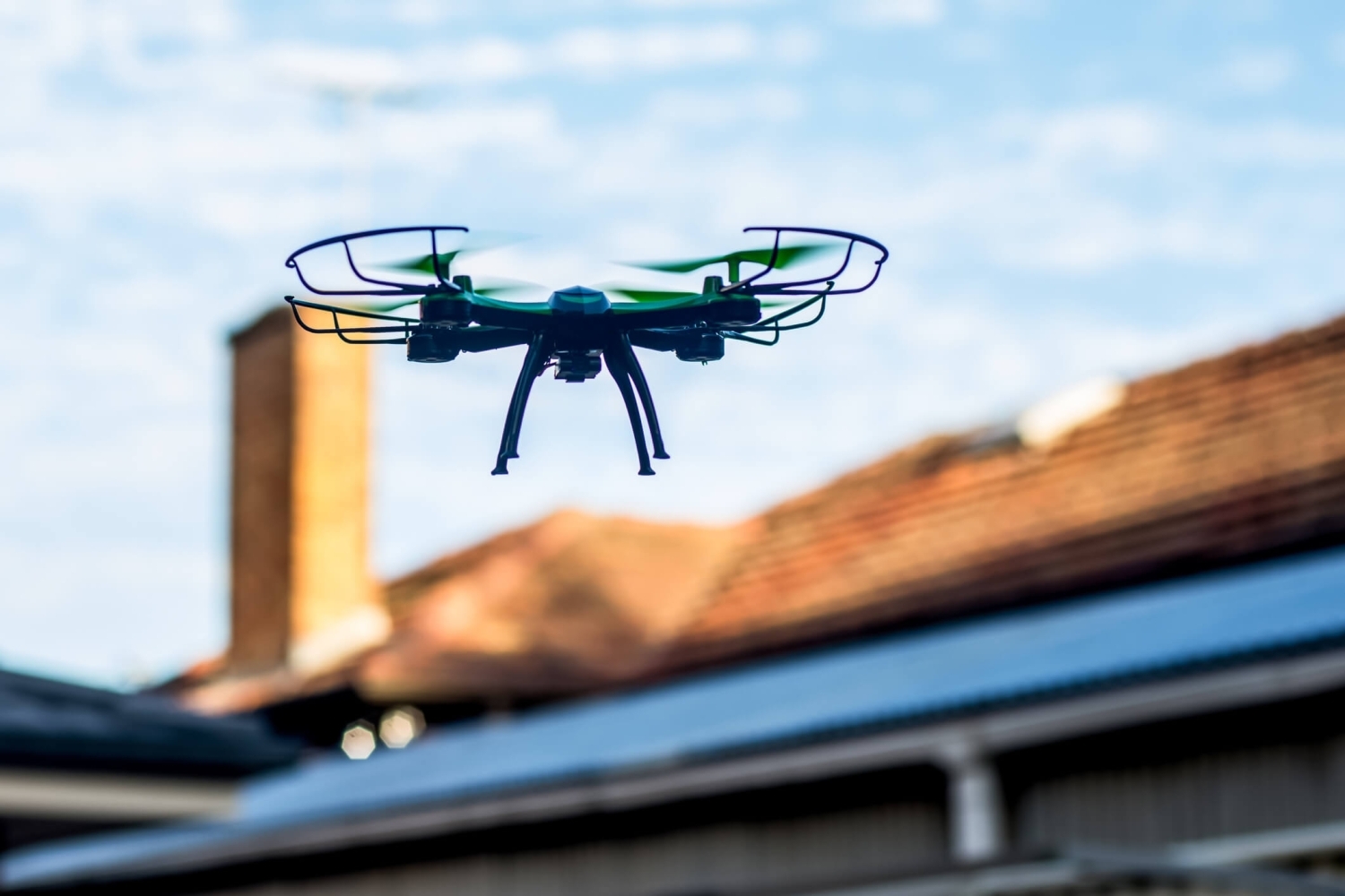 A roofing drone flying over a house with a shingle roof and a chimney in front of a blue sky.